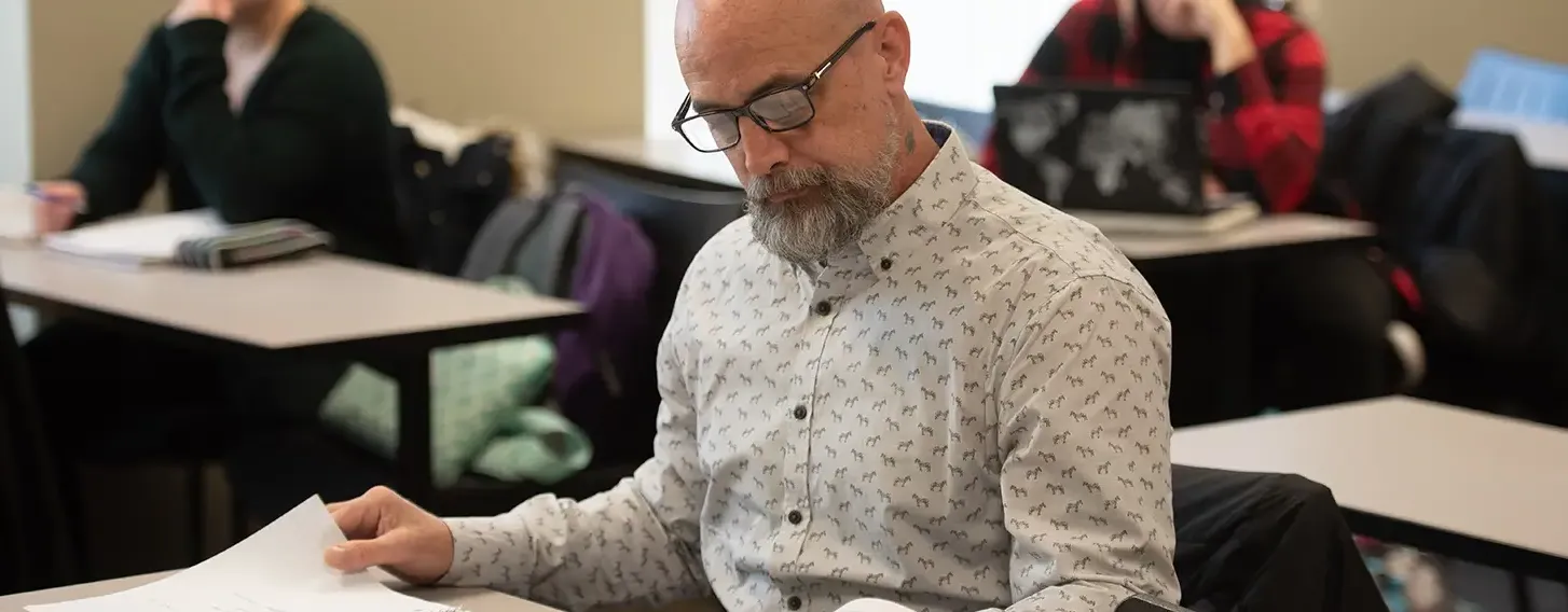 A man with glasses and a beard sitting at a desk with books and papers.